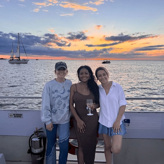 Three people on a boat with a sunset and sailboats in the background. 1