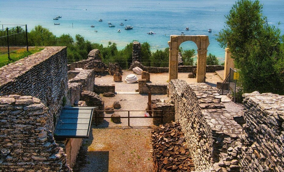 Ancient ruins with an archway, a stone wall, and a pathway leading to the sea. Boats are visible on the water. 4