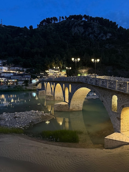 A stone bridge over a calm river at dusk with street lamps and buildings in the background. 5