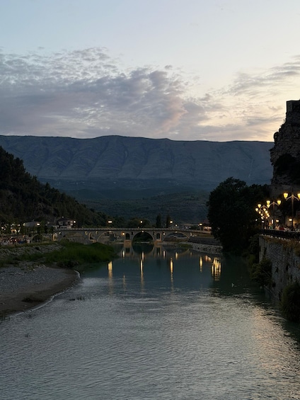 A river with a stone bridge, a calm water surface reflecting lights, and mountains in the background. 2