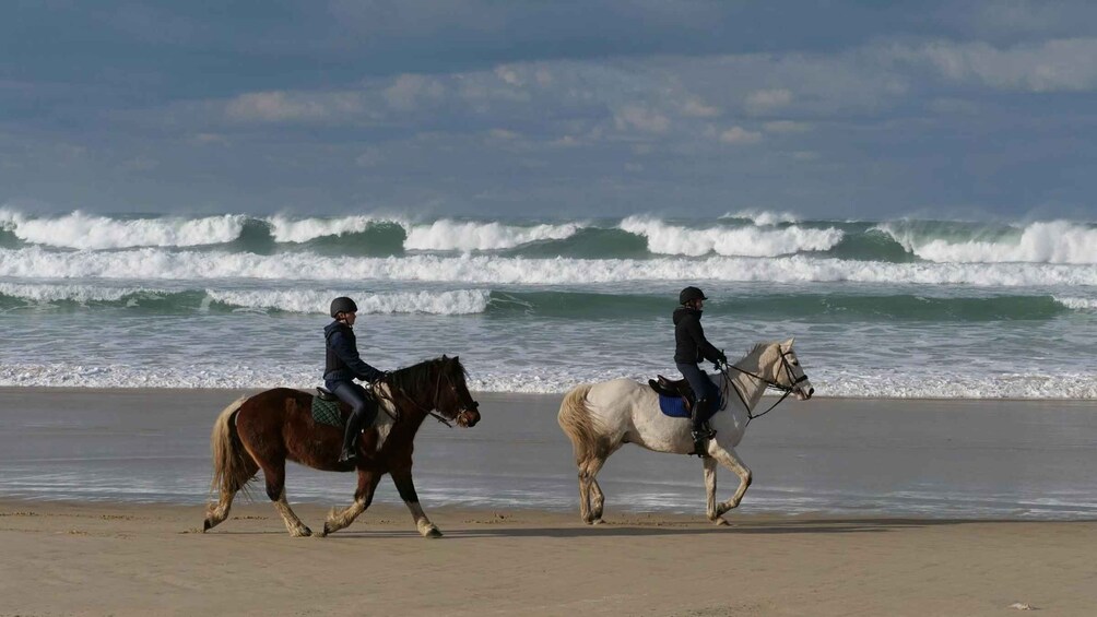Two riders on horses along a beach with waves in the background. 1