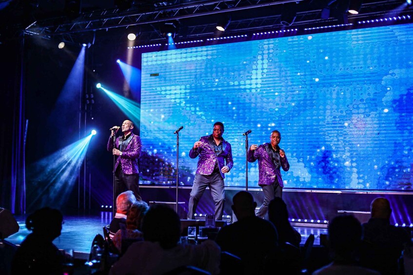 A stage with three performers in sparkling outfits, a large LED screen in the background, and an audience seated in front. 2