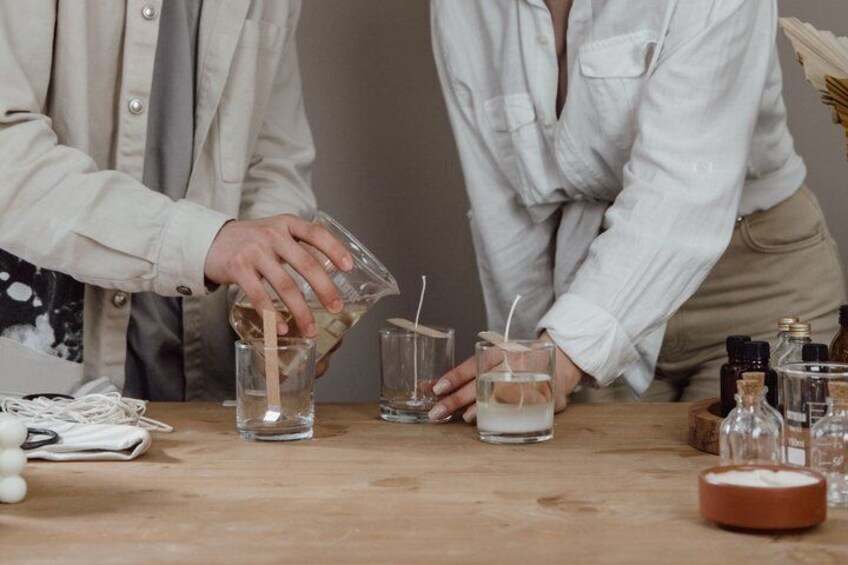 Two people pouring liquids into glasses on a wooden table with various bottles and a candle. 2