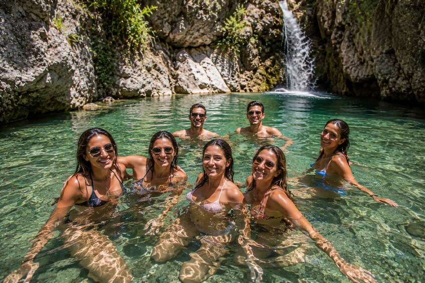 A group of people swimming in a clear, rocky pool with a waterfall in the background. 3