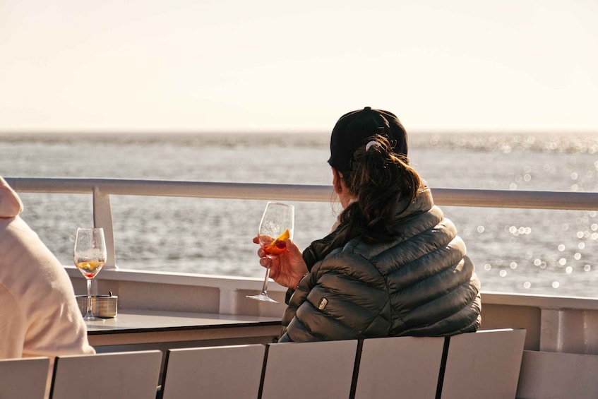 A person sitting by a window on a boat, holding a glass with a drink. 1