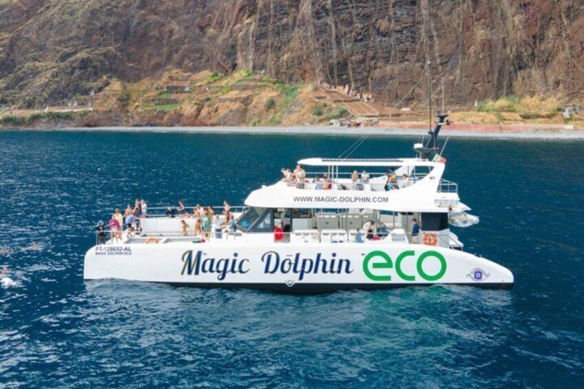 A white boat named 'Magic Dolphin ECO' with people on board, sailing on water with a rocky coastline in the background. 5