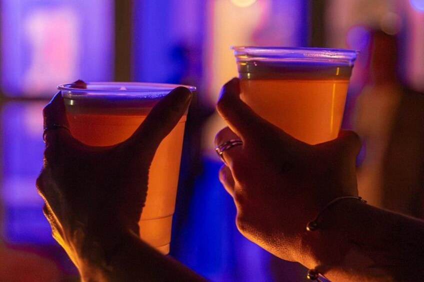 Two hands holding clear plastic cups with amber-colored drinks, in a dimly lit environment with purple lighting. 4