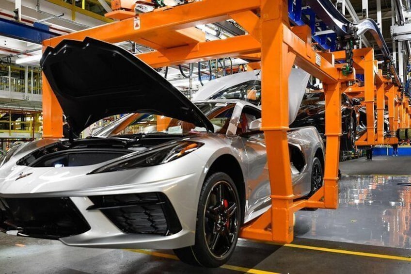 A white sports car with its hood open, positioned under an orange assembly line structure in an industrial setting. 5