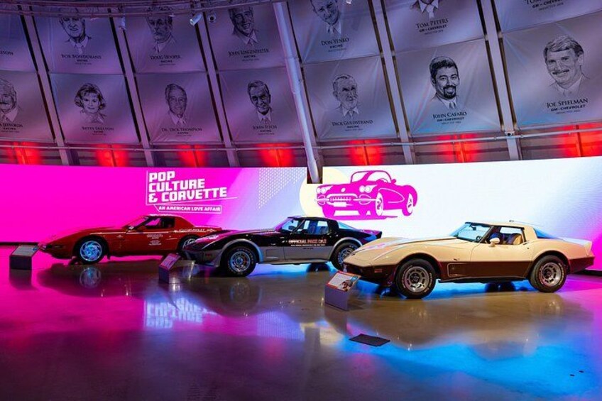 Three classic cars displayed in a museum with a neon sign reading 'POP CULTURE & CORVETTE' and portraits of notable figures on the wall. 3