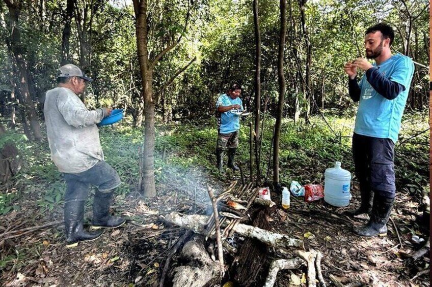 Two people in a forest, one holding a blue object, and a campfire with supplies nearby. 3