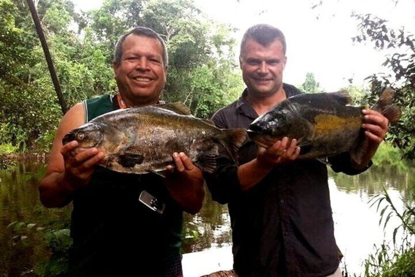 Two individuals holding large fish by a river. 1