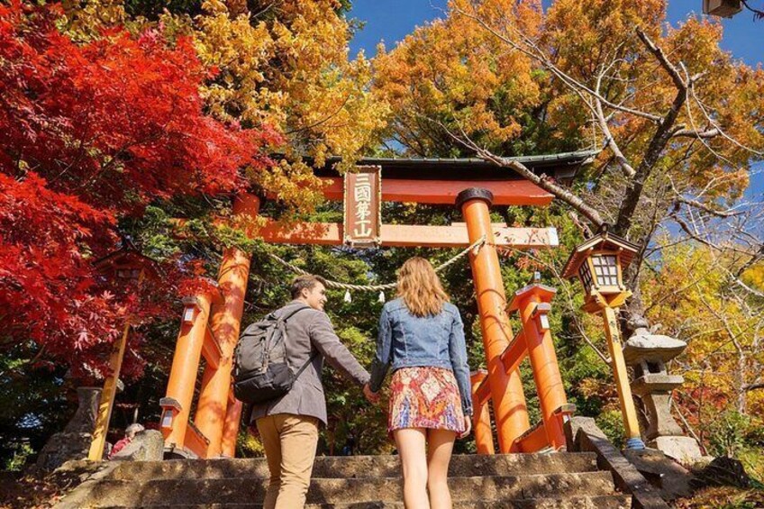 Two people walking up a stone staircase towards a traditional Japanese torii gate surrounded by autumn-colored trees. 3