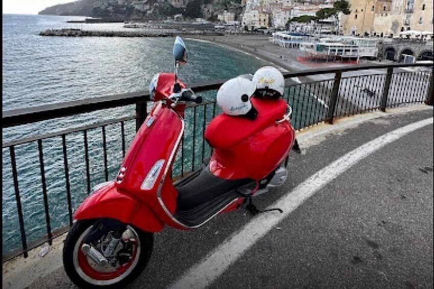 A red scooter parked on a seaside road with a railing, a body of water, and buildings in the background. 2