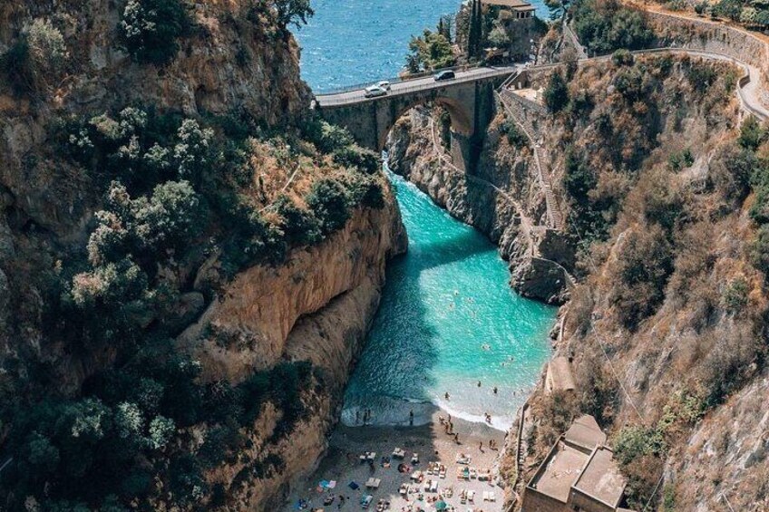 A coastal cliff with a road bridge, turquoise water, and a beach with people. 3