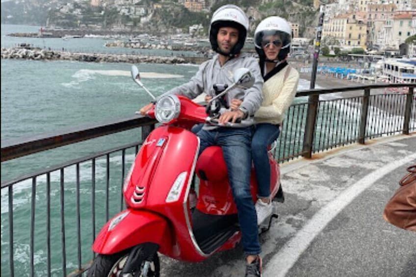 A red scooter with two people wearing helmets, parked by a waterfront with buildings in the background. 1