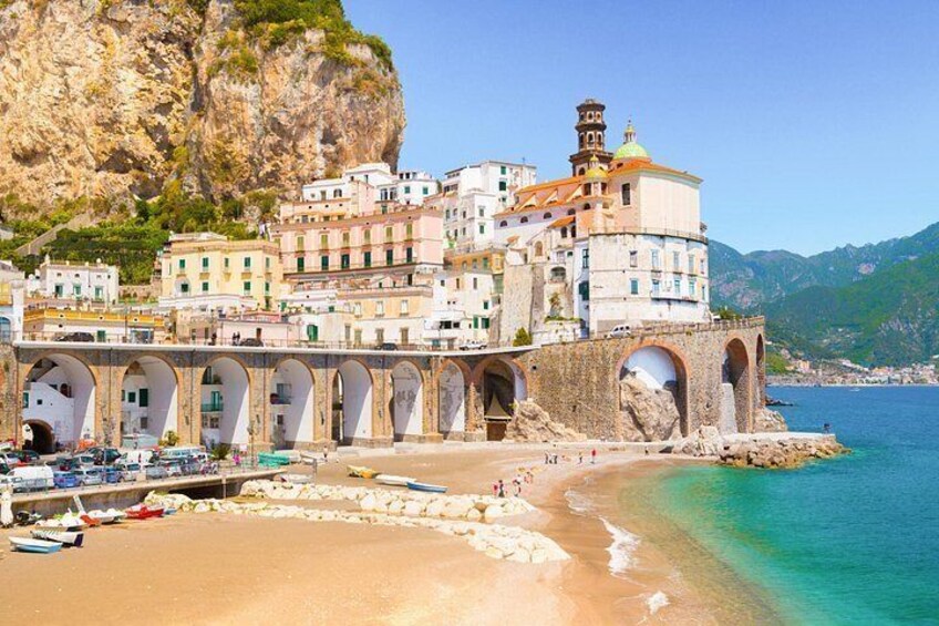 A coastal town with a prominent building featuring arches and a tower, a beach with boats, and a mountainous backdrop. 5