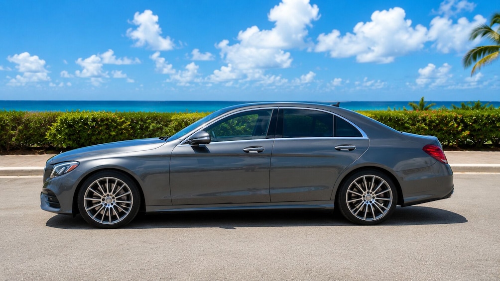 A gray sedan parked on a road with a clear blue sky and ocean in the background. 1