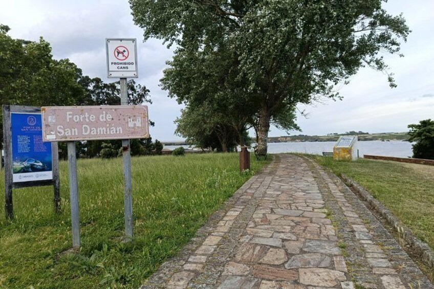 A stone pathway leading to Forte de San Damian, with a no dogs sign and a signboard. 1
