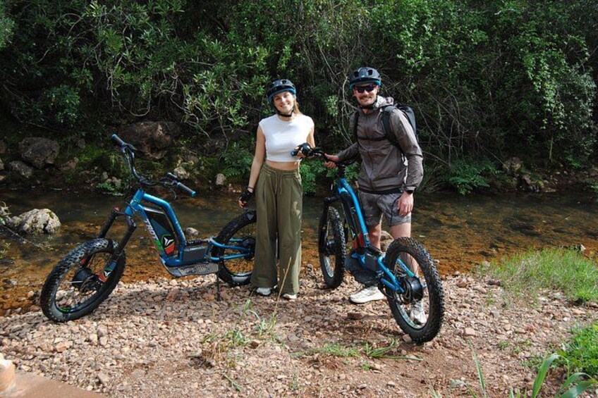 Two people standing by a stream with electric bikes. 2