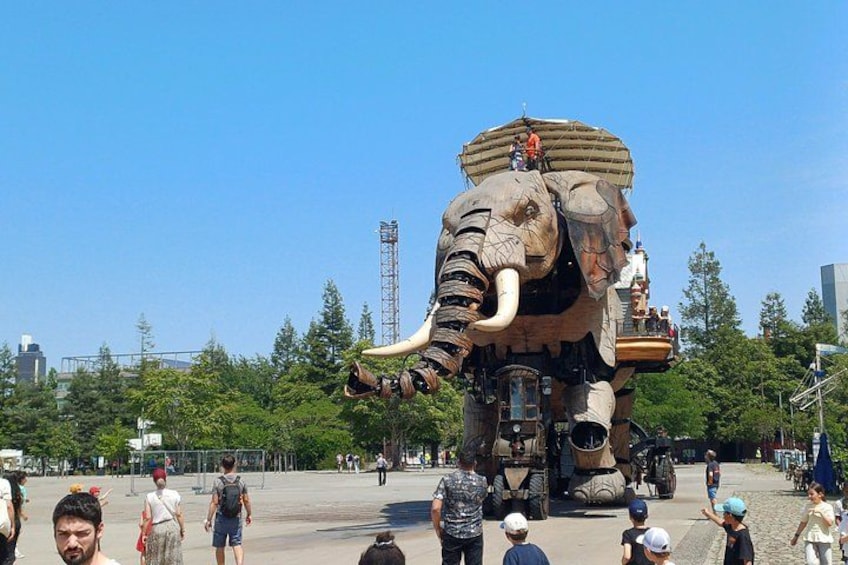 A large elephant statue with a platform on its back, surrounded by people and trees. 1