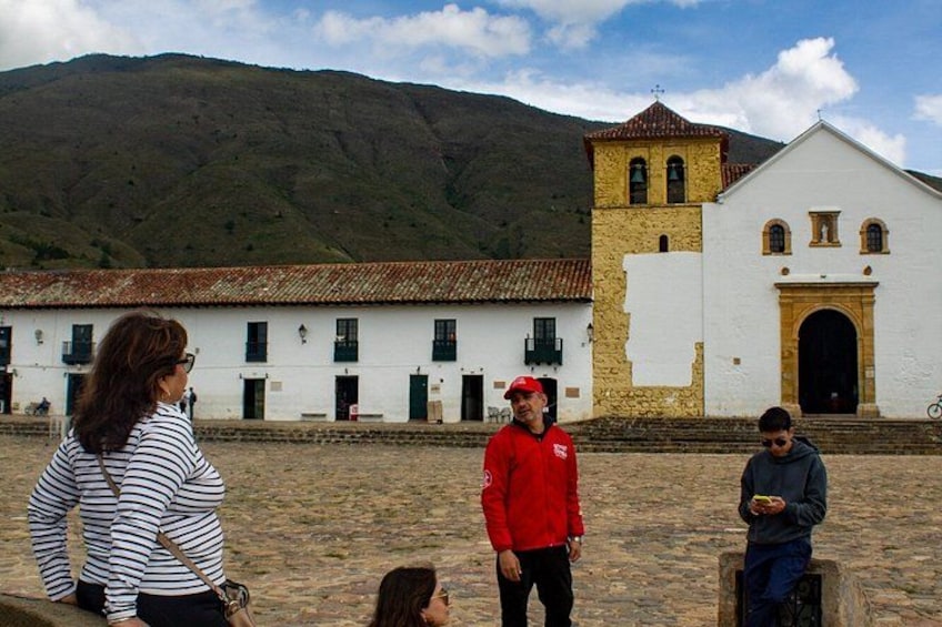 A group of people standing in front of a white building with a bell tower, a mountain in the background, and a cobblestone ground. 3