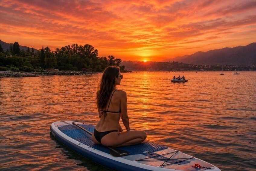 A person sitting on a paddleboard at sunset, with boats and a mountainous landscape in the background. 3