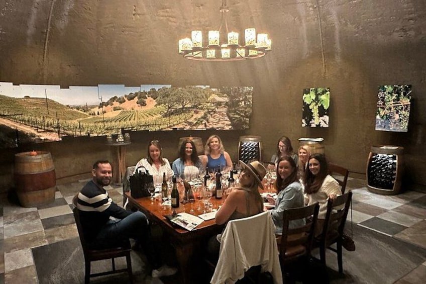 A group of people seated around a table in a rustic interior with a vineyard landscape mural on the wall. 5