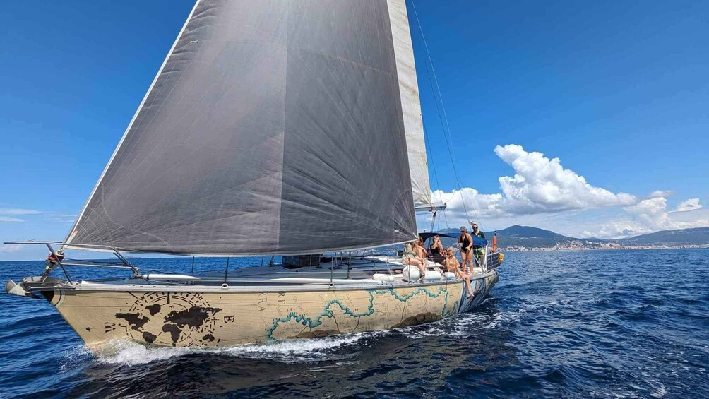 A sailboat with a large sail on the water, a coastline with buildings in the background, and a clear blue sky. 1
