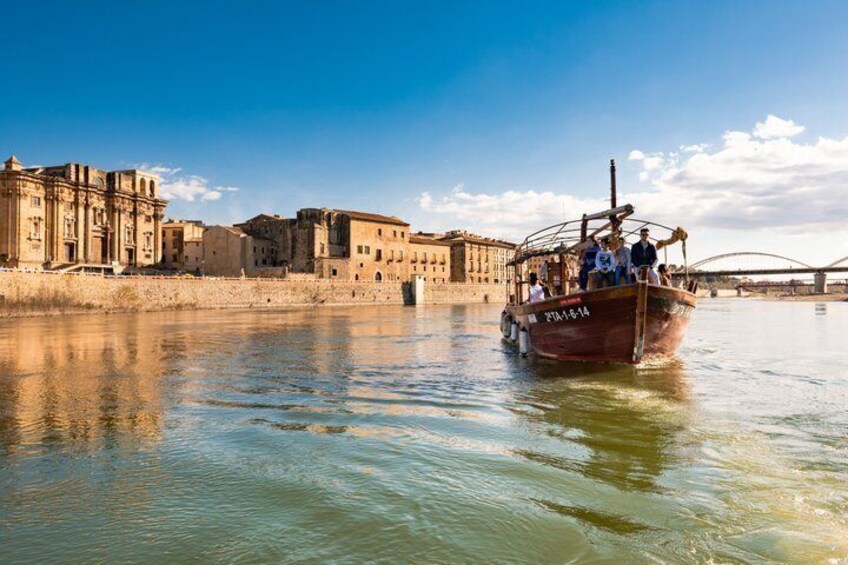 A boat with passengers on a river, a bridge in the background, and historic buildings along the riverbank. 1