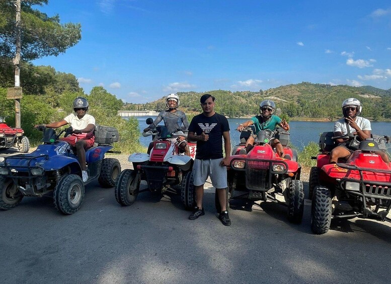 Four people on ATVs parked by a lake with hills in the background. 4