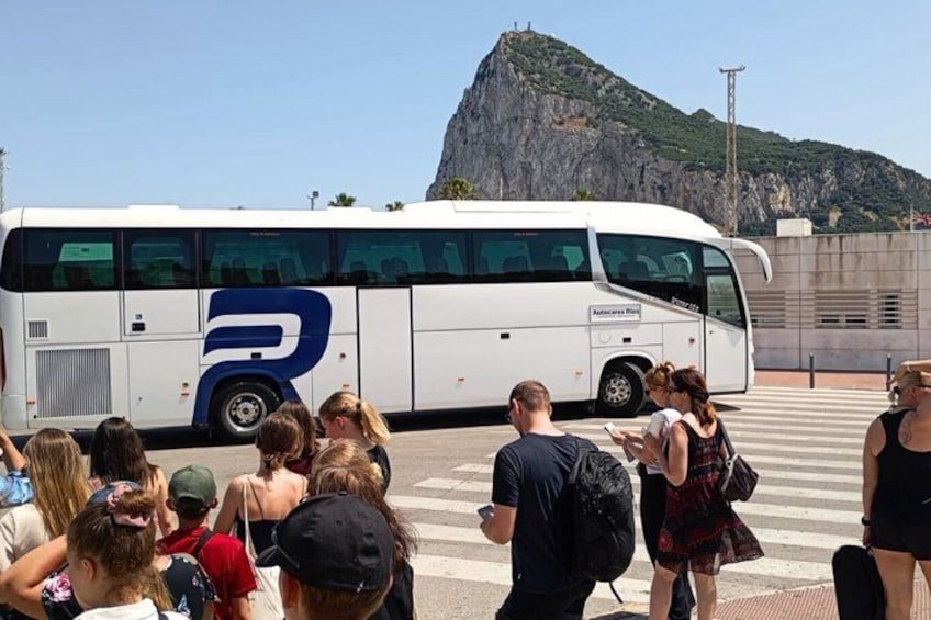 A group of people standing in front of a bus with a large blue 'R' logo, a mountainous landscape in the background, and a clear sky. 3