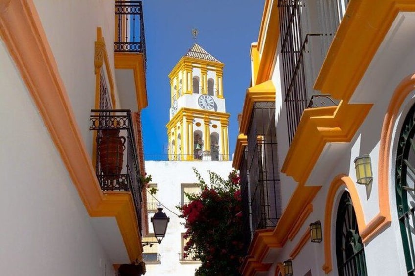 A narrow street with yellow buildings, a clock tower, and a clear blue sky. 4