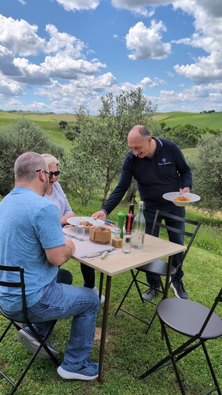 Three people dining outdoors with a table set for a meal, surrounded by greenery and hills in the background. 1