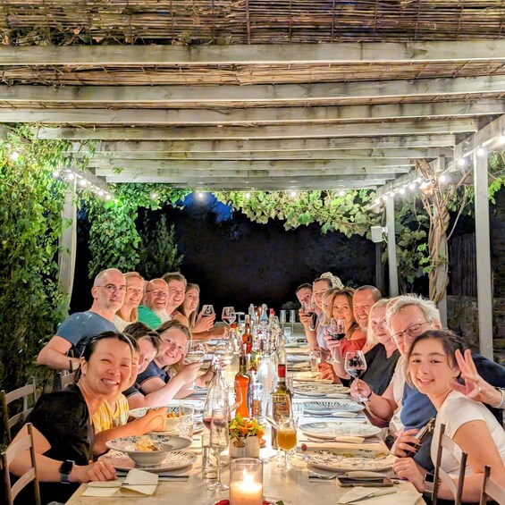 A group of people seated at an outdoor dining table under a pergola with string lights. 2