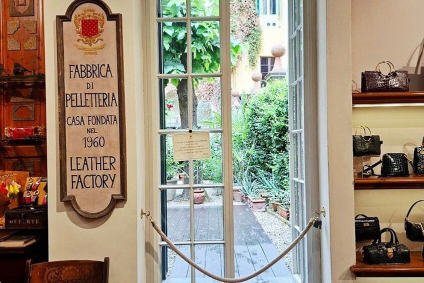 En butik med ett skyltfönster där det står ”FABBRICA DI PELLETERIA CASA FONDATA NEL 1960 LEATHER FACTORY” och med utsikt över en trädgård utanför. 1