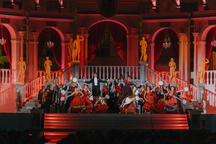 A group of people on a stage with red curtains and statues in a grand building. 2