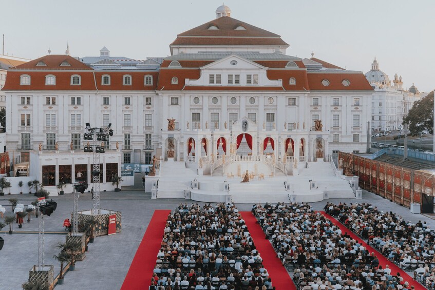 A large white building with red accents, a central stage, and a crowd seated in front. 1