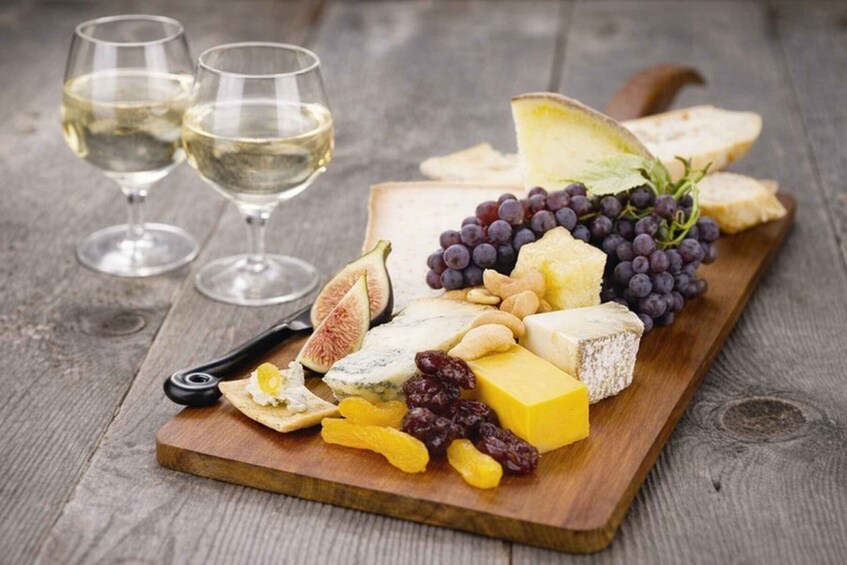 A charcuterie board with cheese, grapes, figs, nuts, and dried fruits, accompanied by two glasses of white wine on a wooden surface. 2