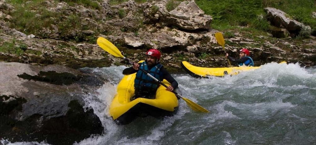 Two people kayaking on a river with rapids, surrounded by rocks and greenery. 1