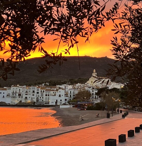 A coastal town at sunset with buildings, a beach, and a hill with a church. 2