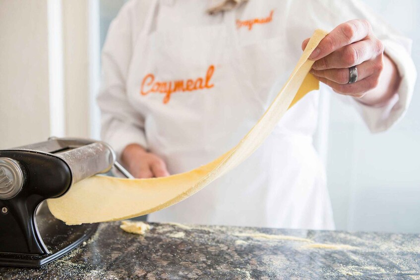 A person in a white apron with 'Cozmeal' written on it, using a pasta machine to roll out dough. 5