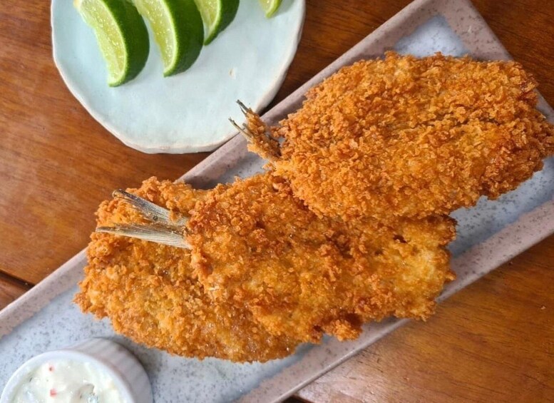 Two breaded and fried fish fillets on a plate with lime wedges and a small bowl of dipping sauce. 5
