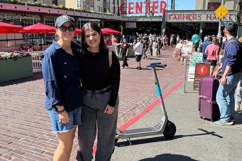 Two people standing in a public square with a scooter and a sign for a Farmers Market in the background. 1