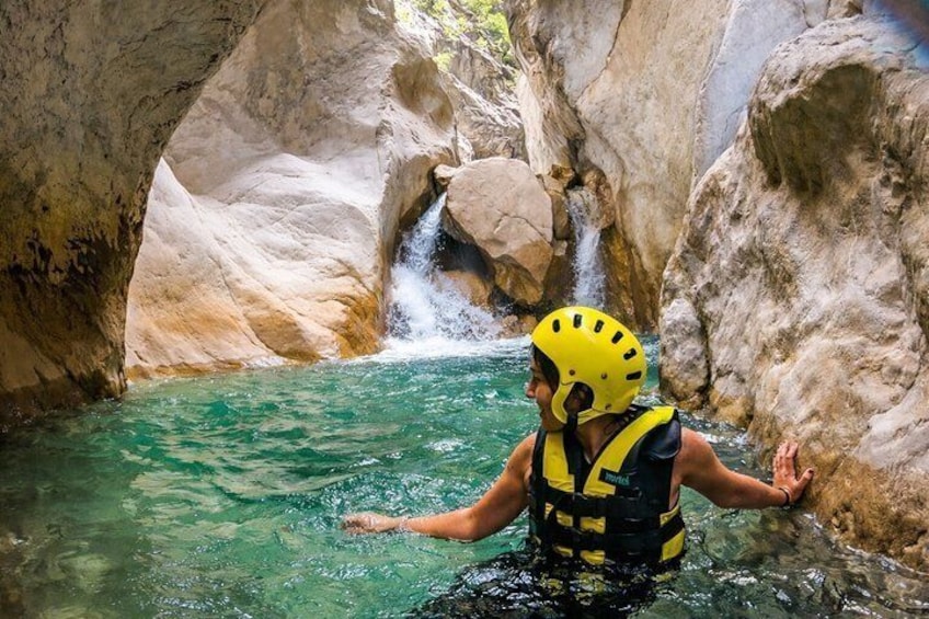 A person in a yellow helmet and life jacket wading through a clear river in a rocky canyon. 2