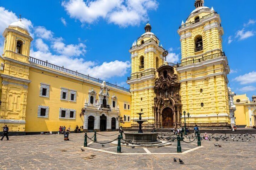 A historic yellow building with two towers, a fountain, and a paved plaza. 2