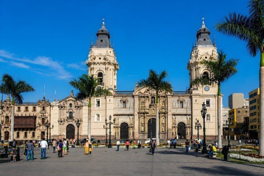 A historic building with two prominent towers and a central dome, surrounded by palm trees and a plaza with people. 5