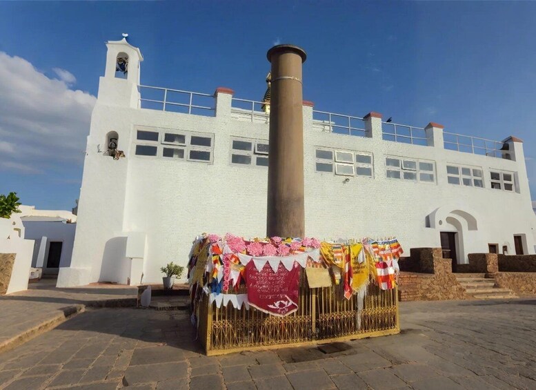 A white building with a bell tower and a large chimney. In front, there's a decorated stall with colorful fabrics and a sign. 2