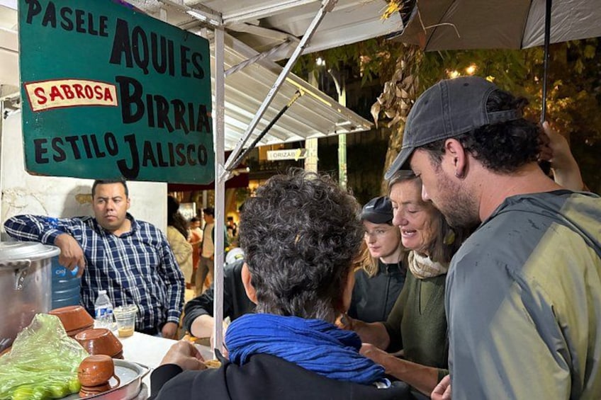 A food stall with a sign indicating it serves 'AQUIES, SABROSA, BIRRIA, ESTILO JALISCO'. 5