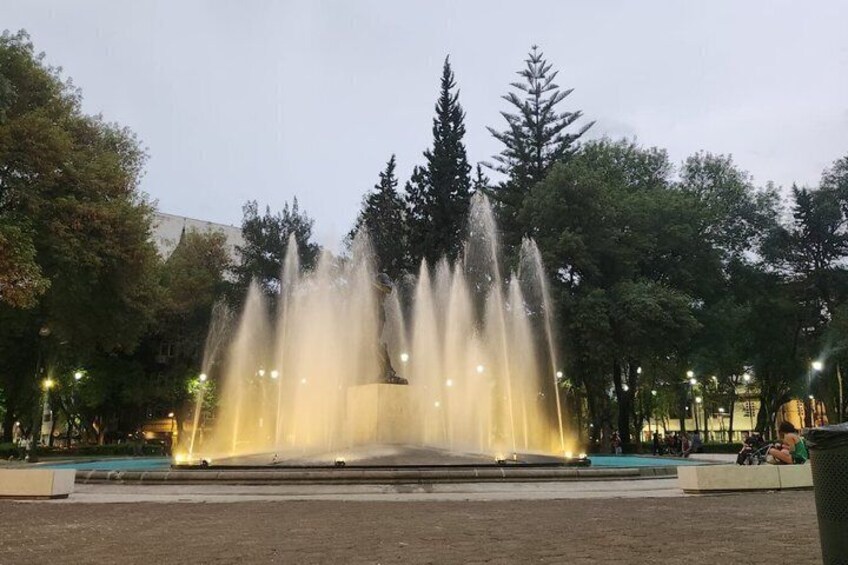 A fountain with water jets in a park with trees and street lamps. 2