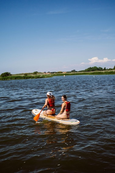 Two people paddleboarding on a calm lake with greenery and houses in the background. 2